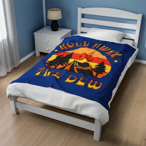 May include: A blue throw blanket with a retro sunset design featuring a tent, trees, and the text "Roll Away The Dew".