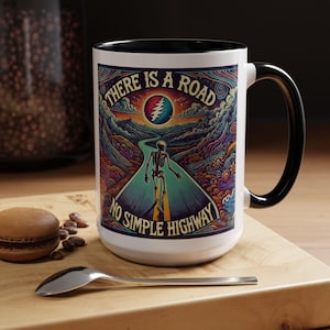 Puede incluir: Taza de cerámica blanca con borde y asa negros. La taza presenta una ilustración colorida de un esqueleto caminando por un camino con el texto "There is a road no simple highway".