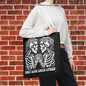 May include: Black tote bag with a white graphic of two skeletons wearing flower crowns and holding hands. The text "They Love Each Other" is printed below the skeletons.