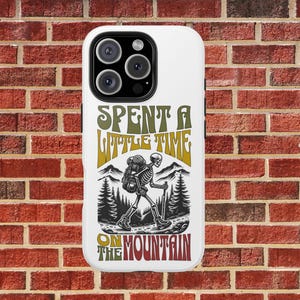 May include: White iPhone case with a skeleton wearing a backpack and hiking in the mountains. The text on the case reads "Spent a little time on the mountain".