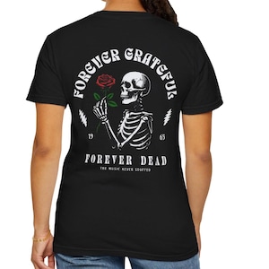 May include: Black t-shirt featuring a white skeleton holding a red rose. The design includes the text "FOREVER GRATEFUL" in a circular arc, with "FOREVER DEAD" below. The phrase "THE MUSIC NEVER STOPPED" is also included.