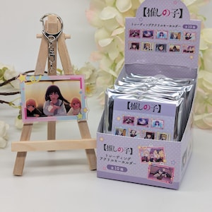 May include: A wooden easel displays a keychain with a rectangular acrylic charm featuring anime characters. A box of similar keychains is behind it. The box and keychain charm have Japanese text. The box is light purple with character images.