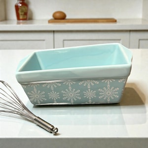 May include: A rectangular, light blue ceramic loaf pan with white snowflake designs around the base. A stainless steel whisk is in the foreground. The pan is on a white countertop, suggesting a kitchen setting.
