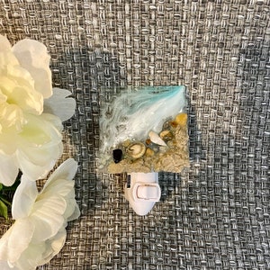 May include: A square night light featuring a beach scene with sand, shells, and a wave design in resin. The night light has a white base with an on/off switch. The background is a gray woven fabric.