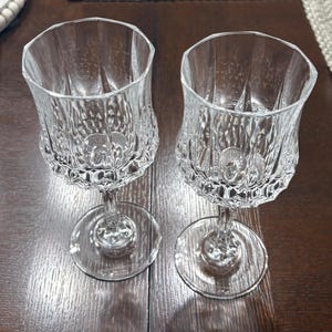 May include: Two clear crystal wine glasses with intricate, textured designs. The glasses have a rounded bowl shape and sit on a stem with a circular base. The glasses are placed on a dark wooden surface.