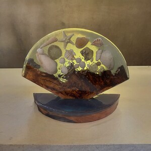 May include: A clear resin lamp base with a wooden stand. The resin contains various seashells and a starfish.