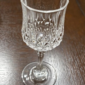 May include: A clear, cut crystal wine glass with a diamond pattern on the bowl. The glass has a stem and a round base. The glass is sitting on a wooden surface.