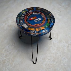 May include: A round side table with a blue resin top embedded with toy cars. The table has a red ring in the center with more toy cars. The table is supported by three black hairpin legs.