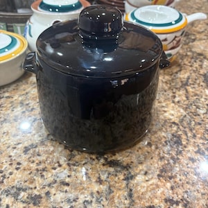 May include: A black ceramic pot with a lid, featuring small handles on the sides. The pot has a glossy finish. Other ceramic bowls and plates are visible in the background, suggesting a kitchenware theme.