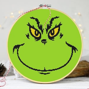 Puede incluir: Bordado de punto de cruz verde de la cara del Grinch con una sonrisa traviesa y ojos amarillos. El bordado está enmarcado en un aro de madera.