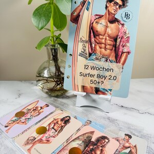 Könnte beinhalten: Eine hellblaue Planerkarte mit einer Illustration eines Mannes mit Surfbrett. Auf der Karte steht der Text "12 Wochen Surfer Boy 2.0 50+?". Mehrere kleinere Karten mit ähnlichen Illustrationen sind sichtbar.