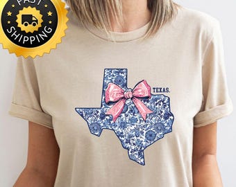 May include: A sand-coloured t-shirt with a blue and white floral Texas outline and a pink bow. The word "Texas" is printed above the state. A gold "Fast Shipping" sticker is in the upper left corner.