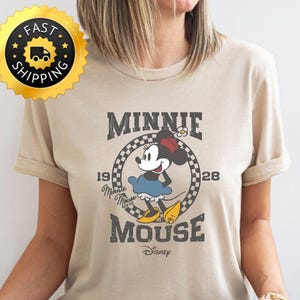 May include: A sand-colored t-shirt featuring a vintage-style graphic of Minnie Mouse. The design includes the text "MINNIE MOUSE" and "1928" with a checkered circle around Minnie. A gold "FAST SHIPPING" sticker is in the upper left.