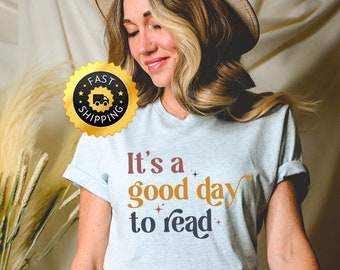 May include: Pale blue t-shirt featuring the phrase "It's a good day to read" in brown, orange, and navy. The shirt is worn with blue denim jeans.