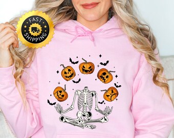 Peut inclure: Sweat à capuche rose pâle sur le thème d'Halloween, avec un squelette méditant, entouré de citrouilles et de chauves-souris. Le sweat à capuche a une poche avant et une capuche à cordon. Le mot "PINK" est en bas de l'image.