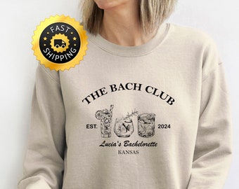 Può includere: Una felpa beige con il testo "The Bach Club Est. 2024" e una grafica di tre bicchieri da cocktail. Il testo "Lucia's Bachelorette Kansas" è sotto la grafica.