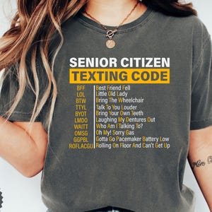 以下が含まれることがあります： 白と黄色で「SENIOR CITIZEN TEXTING CODE」と書かれた濃い灰色のTシャツ。Tシャツには、「Little Old Lady」の「LOL」など、テキストメッセージの頭字語とその意味が記載されています。