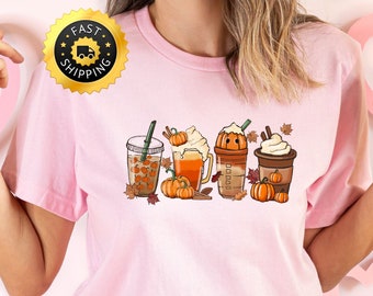 May include: Pink t-shirt featuring a graphic of four autumn-themed drinks: a pumpkin spice latte, a pumpkin beer, a pumpkin iced coffee, and a pumpkin spice tea. The graphic is in a cartoon style and includes pumpkins and autumn leaves.