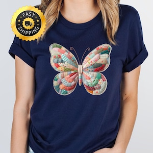 May include: A navy blue t-shirt with a colourful butterfly design. The butterfly has a patchwork quilt pattern in red, green, blue, and yellow. The shirt has short sleeves and a round neckline. A gold sticker with "Fast Shipping" is in the upper left corner.