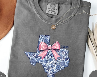 May include: A grey Comfort Colors t-shirt featuring a Texas outline design. The Texas shape is filled with a blue floral pattern and a pink bow. A silver necklace with a pendant is on the shirt.