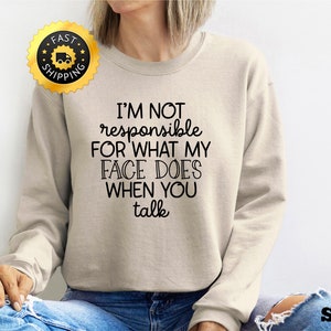 Puede incluir: Una sudadera beige con el texto "No soy responsable de lo que mi cara hace cuando hablas" impreso en negro.