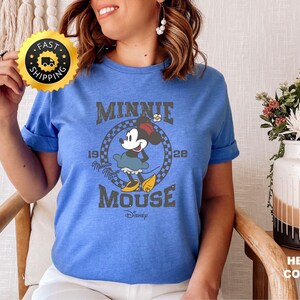 May include: A blue t-shirt featuring a vintage-style graphic of Minnie Mouse. The design includes the text "MINNIE MOUSE" and the year "1928". The shirt is a heather columbia blue color.