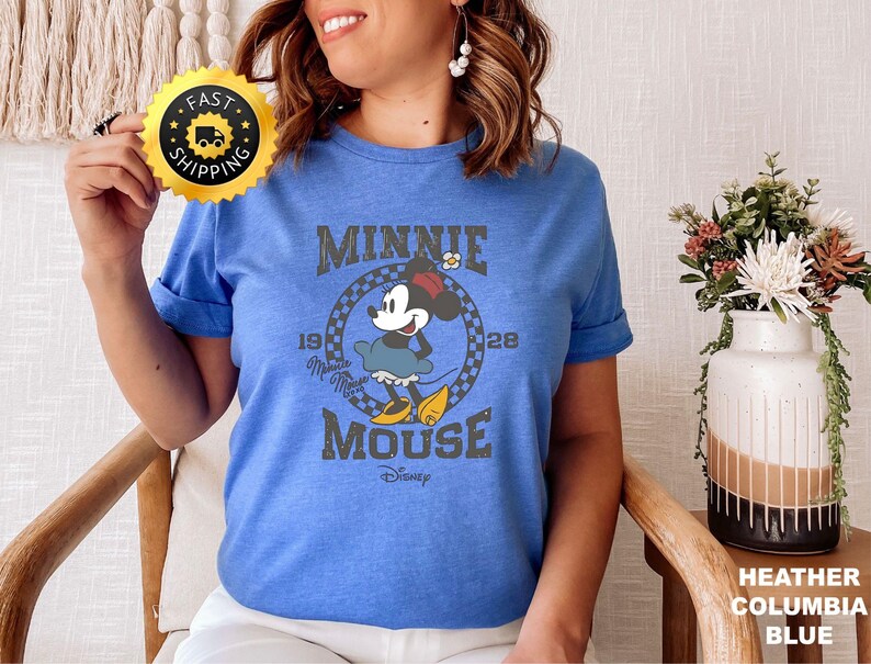 May include: A blue t-shirt featuring a vintage-style graphic of Minnie Mouse. The design includes the text "MINNIE MOUSE" and the year "1928". The shirt is a heather columbia blue color.
