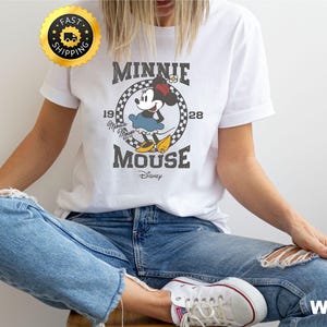 May include: White t-shirt featuring a vintage-style Minnie Mouse graphic. The design includes the text "MINNIE MOUSE" and "1928" in a circular frame. The shirt is paired with blue jeans and white sneakers.