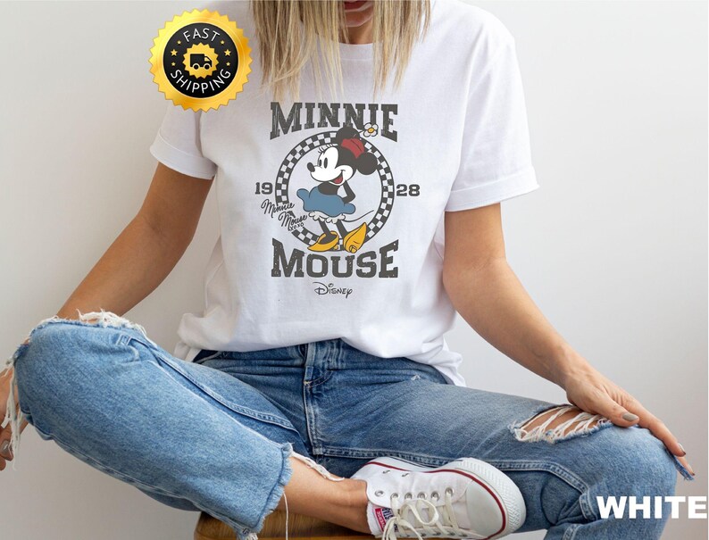 May include: White t-shirt featuring a vintage-style Minnie Mouse graphic. The design includes the text "MINNIE MOUSE" and "1928" in a circular frame. The shirt is paired with blue jeans and white sneakers.