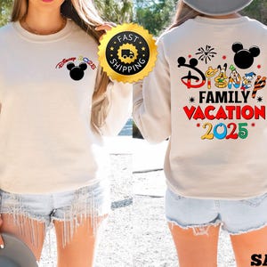 Puede incluir: Una sudadera beige con un gr&aacute;fico de Mickey Mouse en el pecho izquierdo y un gr&aacute;fico m&aacute;s grande en la espalda que dice "Disney Family Vacation 2025" con varios personajes de Disney y fuegos artificiales.