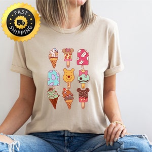 May include: A beige t-shirt with a graphic of various ice cream pops and cones featuring Disney characters. The ice cream pops and cones are in various colors, including pink, blue, green, yellow, and brown. The characters include Mickey Mouse, Minnie Mouse, Winnie the Pooh, and Stitch.