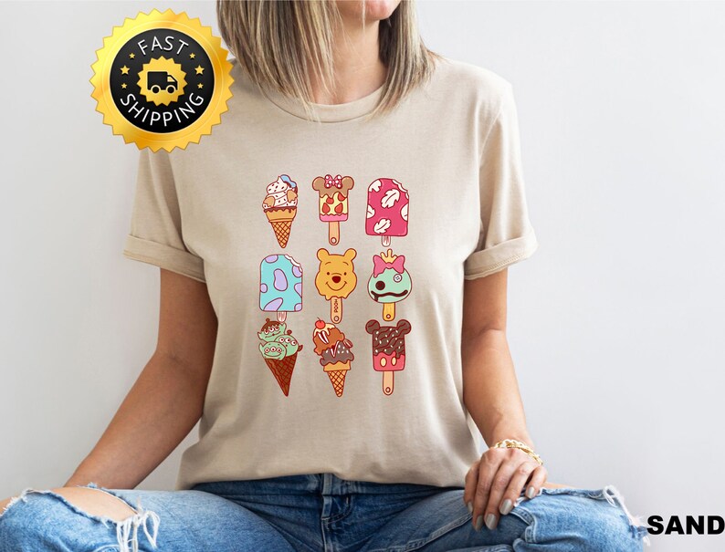 May include: A beige t-shirt with a graphic of various ice cream pops and cones featuring Disney characters. The ice cream pops and cones are in various colors, including pink, blue, green, yellow, and brown. The characters include Mickey Mouse, Minnie Mouse, Winnie the Pooh, and Stitch.