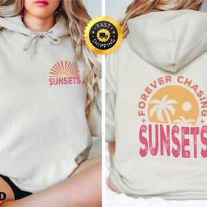 May include: Cream-coloured hoodie with a drawstring hood. The front features a sunset graphic with the word "SUNSETS" in pink. The back displays a circular design with palm trees, the sun, and the words "FOREVER CHASING SUNSETS".