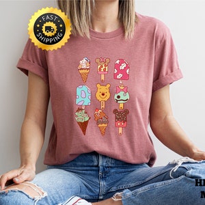 May include: A heather mauve t-shirt with a graphic of various ice cream and popsicle designs featuring Disney characters. The characters include Mickey Mouse, Minnie Mouse, Winnie the Pooh, and a green alien. The text "FAST SHIPPING" is on a gold star sticker in the top left corner of the image.