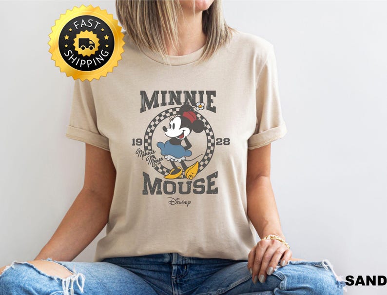 May include: A sand-colored t-shirt featuring a vintage-style graphic of Minnie Mouse. The design includes the text "MINNIE MOUSE" and "1928" with a checkered circle around Minnie. A gold "FAST SHIPPING" sticker is in the upper left.