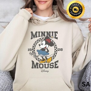 May include: Sand-colored hoodie featuring a graphic of Minnie Mouse in a vintage style. The design includes the text "MINNIE MOUSE" and "1928" with a checkered circle around Minnie. The Disney logo is at the bottom.