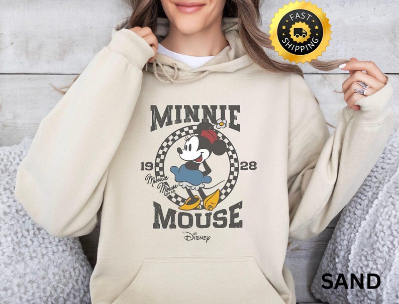 May include: Sand-colored hoodie featuring a graphic of Minnie Mouse in a vintage style. The design includes the text "MINNIE MOUSE" and "1928" with a checkered circle around Minnie. The Disney logo is at the bottom.