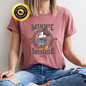 May include: Mauve-colored t-shirt featuring a vintage-style graphic of Minnie Mouse. The design includes the text "MINNIE MOUSE" and "1928" in a circular frame. The shirt is made of soft material and is suitable for casual wear.