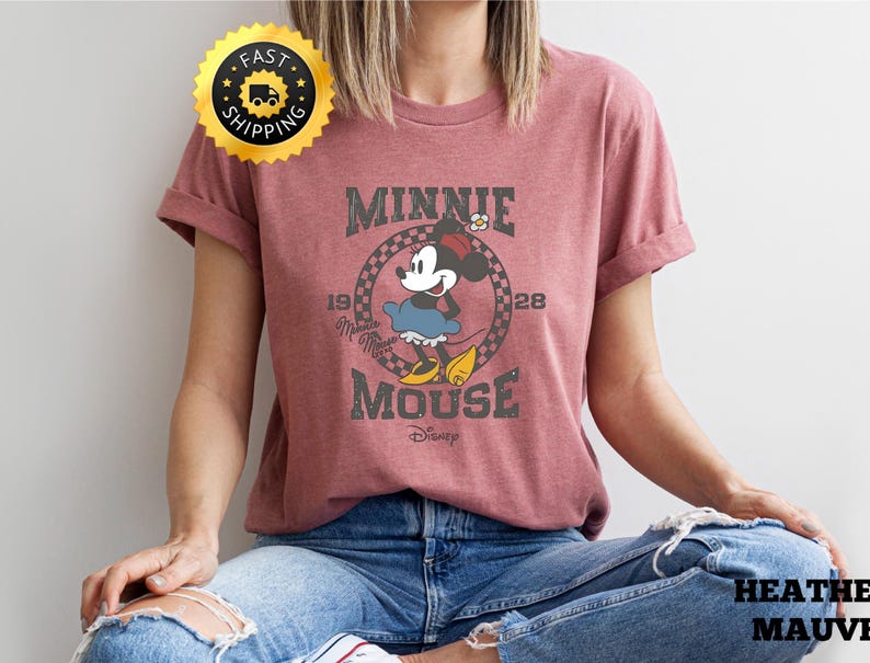 May include: Mauve-colored t-shirt featuring a vintage-style graphic of Minnie Mouse. The design includes the text "MINNIE MOUSE" and "1928" in a circular frame. The shirt is made of soft material and is suitable for casual wear.