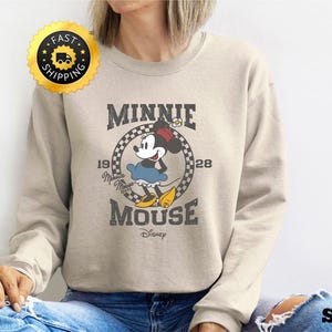 May include: Sand-colored sweatshirt featuring a vintage Minnie Mouse graphic. The design includes Minnie in a blue dress and yellow shoes, with the text "MINNIE MOUSE 1928 Disney". The sweatshirt has a crew neck and long sleeves.