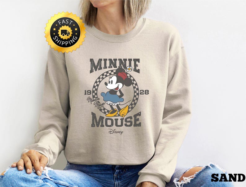 May include: Sand-colored sweatshirt featuring a vintage Minnie Mouse graphic. The design includes Minnie in a blue dress and yellow shoes, with the text "MINNIE MOUSE 1928 Disney". The sweatshirt has a crew neck and long sleeves.