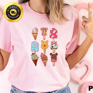 May include: Pink t-shirt with a graphic of various cartoon characters on popsicles and ice cream cones. The characters include Mickey Mouse, Minnie Mouse, Winnie the Pooh, and others.