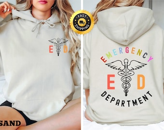 Peut inclure: Un sweat à capuche crème avec un symbole médical noir et blanc et le texte "ED" sur le devant. Le dos du sweat à capuche porte le texte "EMERGENCY DEPARTMENT" en couleurs arc-en-ciel autour du symbole médical.
