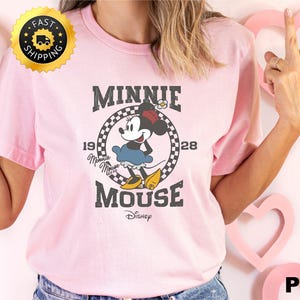 May include: A light pink t-shirt featuring a graphic of Minnie Mouse. The design includes the text "MINNIE MOUSE" above and below the character, with "1928" on either side. The Disney logo is at the bottom.