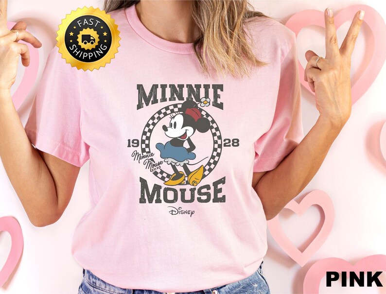 May include: A light pink t-shirt featuring a graphic of Minnie Mouse. The design includes the text "MINNIE MOUSE" above and below the character, with "1928" on either side. The Disney logo is at the bottom.