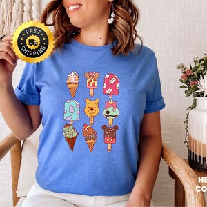 May include: A blue t-shirt with a graphic of various ice cream pops featuring Disney characters like Winnie the Pooh, Mickey Mouse, and Stitch. The ice cream pops are arranged in a grid pattern on the shirt.