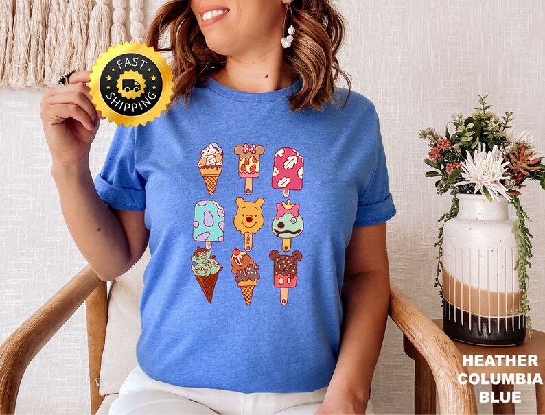 May include: A blue t-shirt with a graphic of various ice cream pops featuring Disney characters like Winnie the Pooh, Mickey Mouse, and Stitch. The ice cream pops are arranged in a grid pattern on the shirt.