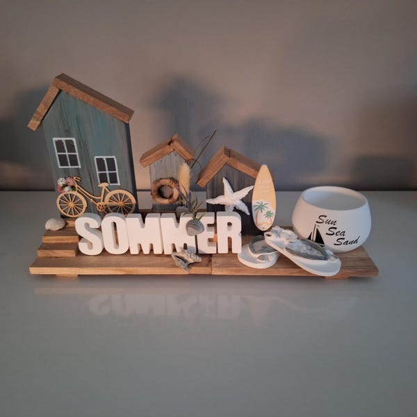 May include: A wooden summer-themed decorative display. It features miniature beach huts, a bicycle, a surfboard, a starfish, and the word "SOMMER" in white letters. A white ceramic pot with the words "Sun Sea Sand" is also included.