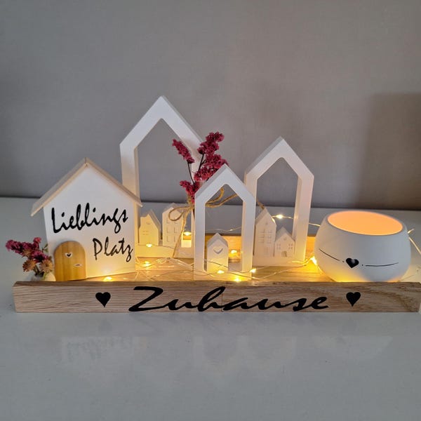 May include: A wooden tray with a white house cutout that says "Lieblings Platz" and a white ceramic candle holder with a heart design. The tray also has a string of fairy lights and the words "Zuhause" written on it.