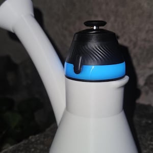 May include: A white water pipe with a curved neck and a wide base. The top features a black cap with a textured, spiraled design and a bright blue ring accent. The overall structure is smooth and matte, with a functional spout extending upward.